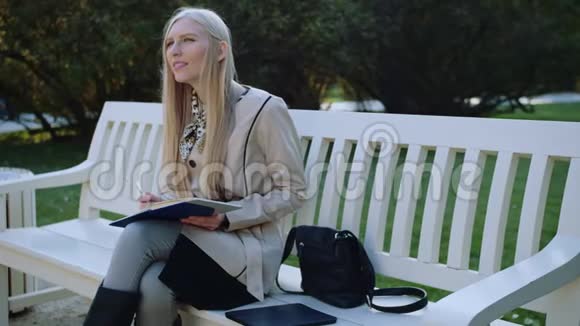 美丽的女人坐在秋天公园的长椅上在笔记本上做笔记视频的预览图