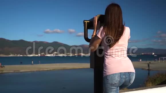 穿着粉红色衬衫和蓝色牛仔裤的旅行者年轻女子透过硬币固定望远镜观看视频的预览图