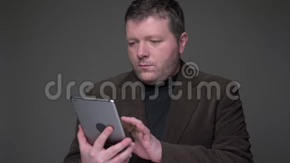 帅气的黑发中年商人穿着西装在平板电脑上冲浪在灰色背景下情绪化地反应视频的预览图