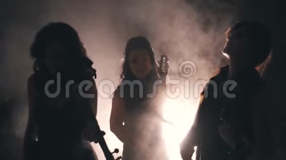 三个伟大的女孩小提琴手在一个烟雾弥漫的房间里演奏光线从烟雾中穿过很漂亮的灯光视频的预览图