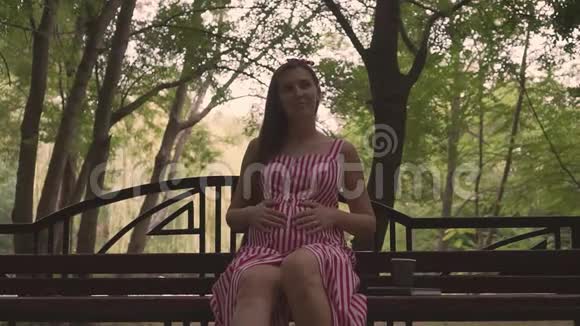 公园里树背景上的怀孕女孩一个穿条纹连衣裙的女孩坐在长凳上抚摸着肚子视频的预览图