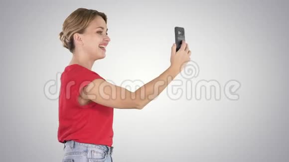 美丽的年轻女子走着拿着智能手机在梯度背景上拍照和自拍视频的预览图