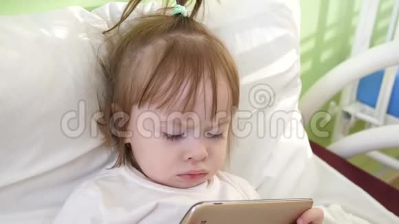 可爱的小女孩躺在儿童医院床上在智能手机上看有趣的卡通片视频的预览图