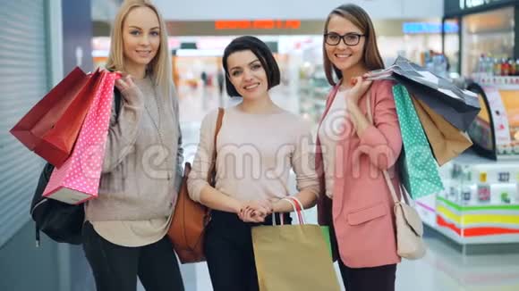 购物中心里站着五颜六色的纸袋看着照相机和照相机的快乐女孩的画像视频的预览图
