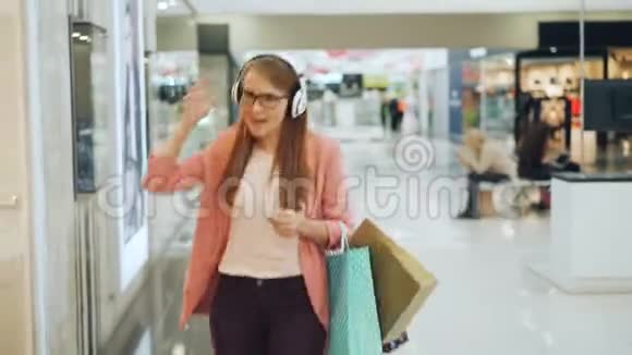 快乐的女人在耳机里享受音乐在购物中心大厅拿着纸袋跳舞唱歌女孩是视频的预览图