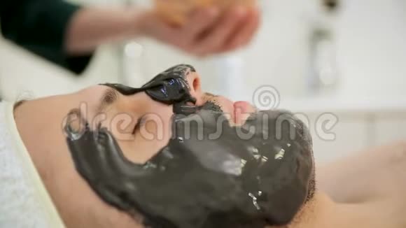 美容师把一个黑色的面具戴在男人的脸上用一个黑色组织的恢复活力的化妆面具戴在脸上视频的预览图