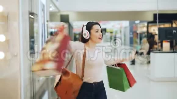 快乐的女购物狂在购物中心里玩得很开心用耳机听音乐用明亮的袋子跳舞和跳舞视频的预览图