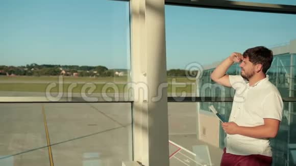 留着胡子的人站在机场的窗边拿着护照等着飞机视频的预览图