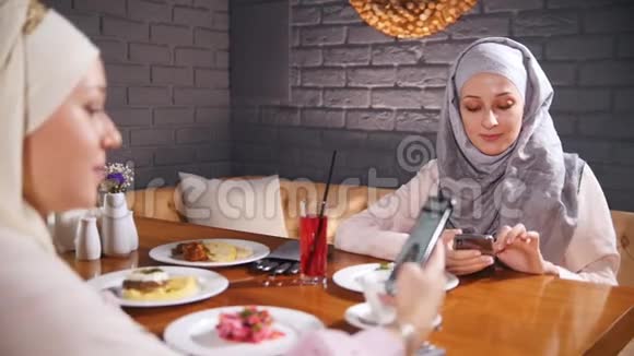两个穆斯林妇女坐在餐厅的一张桌子上坐在电话里在电话里显示照片视频的预览图