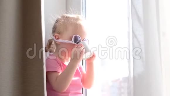 可爱的金发小女孩戴着粉红色太阳镜在窗边喝着早间咖啡或茶看着外面视频的预览图