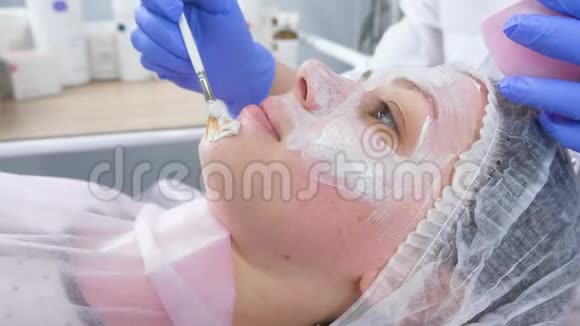美容师用刷子把白色面具戴在女人的脸上戴着蓝色橡胶手套的美容师的手面部视频的预览图