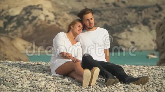 年轻漂亮的夫妇坐在海边温暖的鹅卵石上他们愉快地交流一起度过时光视频的预览图