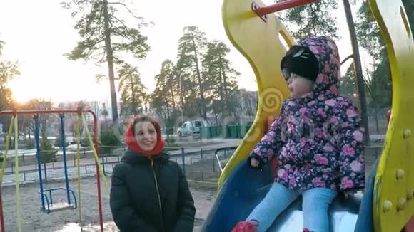 一位年轻漂亮的母亲穿着一件五颜六色的夹克照顾她的女儿她坐在一个高大的春天公园的滑梯上视频的预览图
