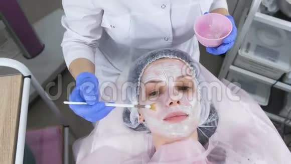 美容师用刷子把白色面具戴在女人的脸上戴着蓝色橡胶手套的美容师的手面部视频的预览图