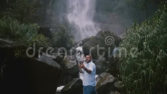 年轻快乐微笑旅游博主男子在史诗般雄伟的斯里兰卡热带丛林瀑布上拍摄智能手机自拍视频的预览图