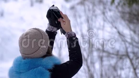 年轻漂亮的女孩走在白雪皑皑的冬林里她高兴一个女孩正在智能手机上拍照做一个视频的预览图