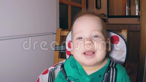 小可爱的没有牙齿的男孩绿色的身体广泛微笑坐在厨房的喂食椅附近的冰箱视频的预览图