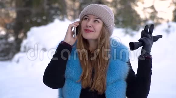 一个年轻漂亮的女孩在一个下雪的冬天森林里打电话她高兴穿着冬帽和毛皮外套的女孩慢慢视频的预览图