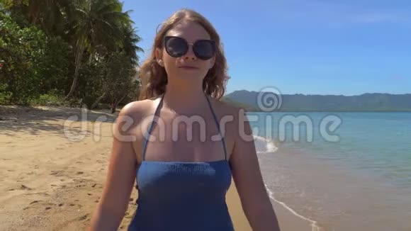 年轻的欧洲女孩戴着太阳镜和蓝色连衣裙在沙滩上散步背景是视频的预览图