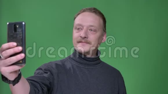 金发白种人的养老金在灰色套头衫在智能手机上的绿色背景拍摄自拍照片视频的预览图