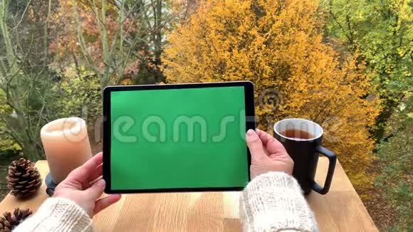 女孩持有平板电脑与预先键好的绿色屏幕黑色杯子与茶在露台上秋黄和橙色的树木概念视频的预览图