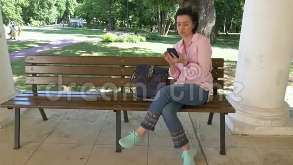 一位坐在长凳上的公园里的黑发女人看着她智能手机上的照片视频的预览图