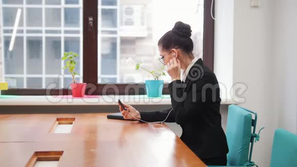 商业概念一个戴眼镜的女人坐在桌子旁把耳机插入耳朵里视频的预览图