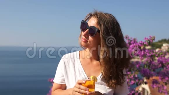 海上海岸一个漂亮女孩的肖像一个女人站在有花的露台上拿着一个玻璃杯视频的预览图