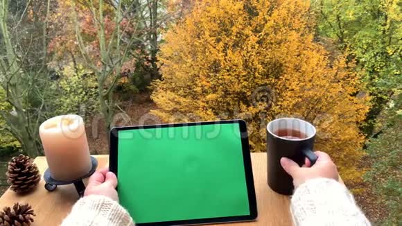 女孩持有平板电脑与预先键好的绿色屏幕黑色杯子与茶在露台上秋黄和橙色的树木概念视频的预览图