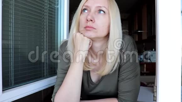 一幅年轻漂亮的金发女子的肖像蓝色的眼睛站在早晨打开的窗户附近看着视频的预览图