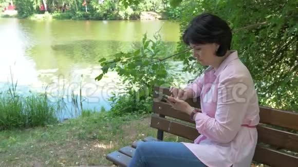 一位坐在长凳上的公园里的黑发女人看着她智能手机上的照片视频的预览图