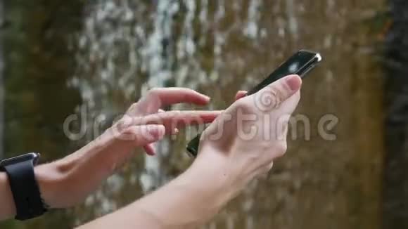有智能手表的女人在瀑布的背景下使用智能手机依赖互联网生态旅游视频的预览图