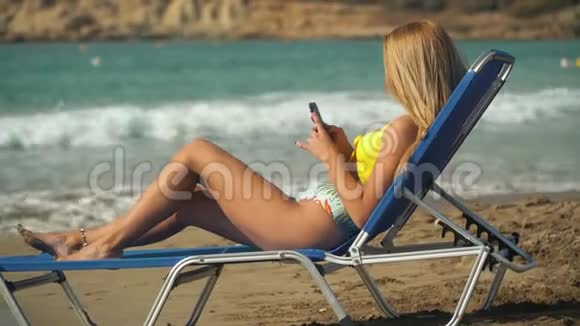 年轻的金发女人在海滩上晒太阳躺在甲板椅上背景是大海女孩在手机上发短信视频的预览图