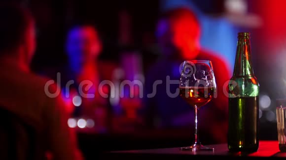 带霓虹灯的酒吧一个年轻人拿着绿色的瓶子和一杯啤酒坐在他的朋友旁边干杯视频的预览图