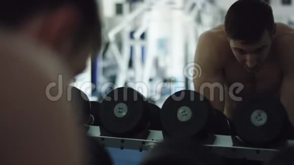 年轻强壮的男人在健身房里对着镜子看哑铃视频的预览图