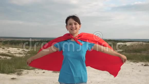 美丽的女超级英雄穿着红色斗篷在蓝天前穿过田野微笑着慢动作少女梦视频的预览图