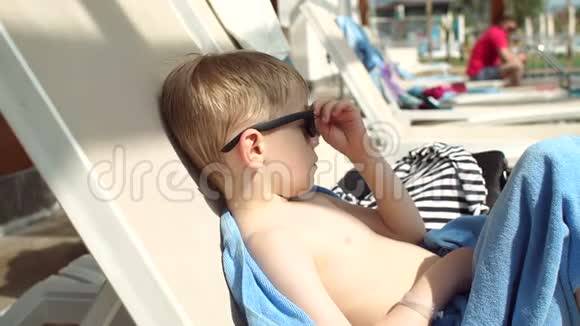 一个金发的小男孩戴着太阳镜在阳光休息室里晒太阳视频的预览图