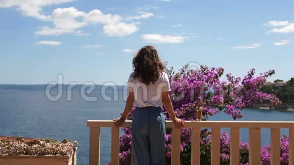 站在海边露台上的长卷发的年轻女子在花丛附近和远处视频的预览图