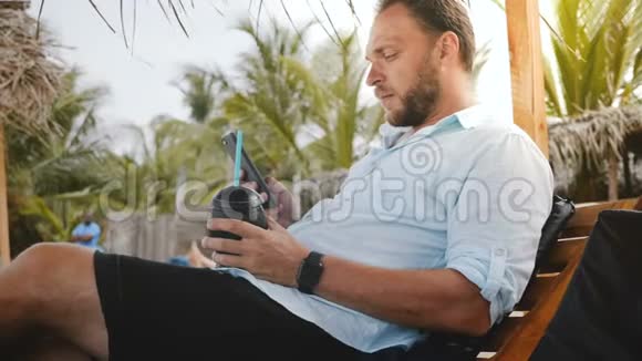 成功的快乐男游客带着智能手表和水果饮料坐在沙滩椅上使用智能手机购物应用程序视频的预览图