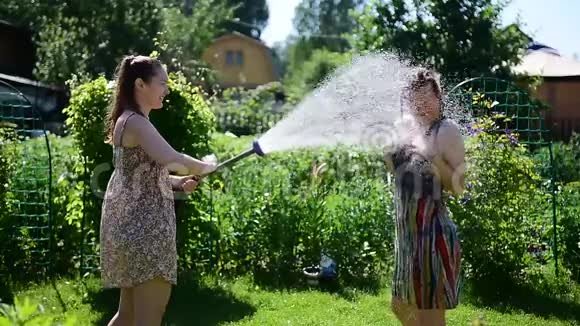在炎热的夏日阳光明媚的日子里可爱的女孩从绿色草坪上的花园软管里倒水穿着礼服的金发美女视频的预览图