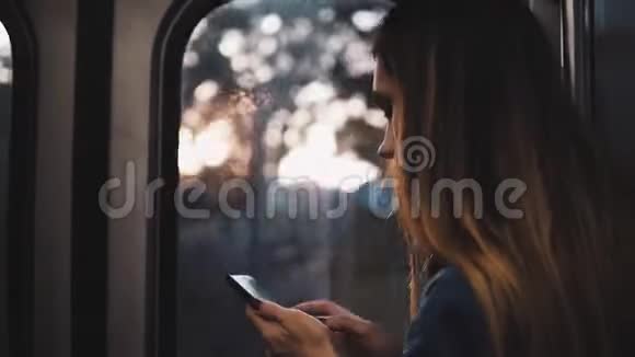 年轻的黑发女人在移动地铁的某个地方日落时女孩用智能手机站在窗户附近视频的预览图