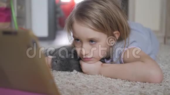 可爱的小女孩拿着数字平板电脑抱着你最喜欢的猫躺在地板上美丽的女孩在玩视频的预览图