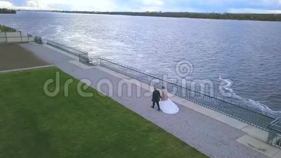 幸福的新婚夫妇在阴云密布的天空背景下在宁静的河边散步和牵手剪辑视频的预览图
