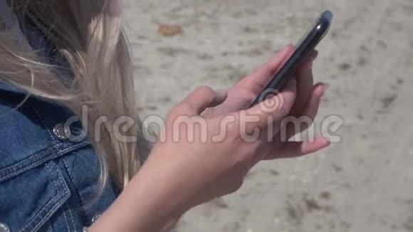 年轻的金发女郎穿着海和海滩背景的牛仔夹克她的头发在智能手机上飘动视频的预览图