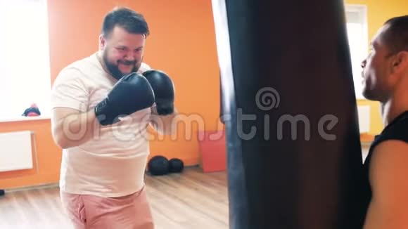 与个人教练的个人训练一个戴拳击手套的胖家伙胜过健身俱乐部的拳击袋重量损失视频的预览图