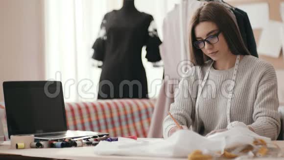女时装设计师在桌子旁的工作室里用一块布一根彩色线和一台笔记本电脑画一幅素描取一个视频的预览图