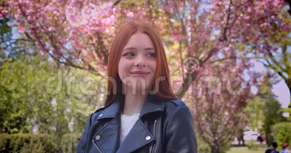 迷人的生姜模型在皮夹克摆在相机在粉红色的花卉花园视频的预览图