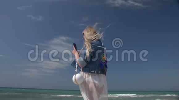 年轻的金发女人手里拿着智能手机背景是大海和蓝天视频的预览图