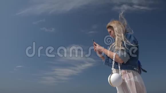 年轻的金发女人手拿智能手机对着蓝天慢镜头视频的预览图