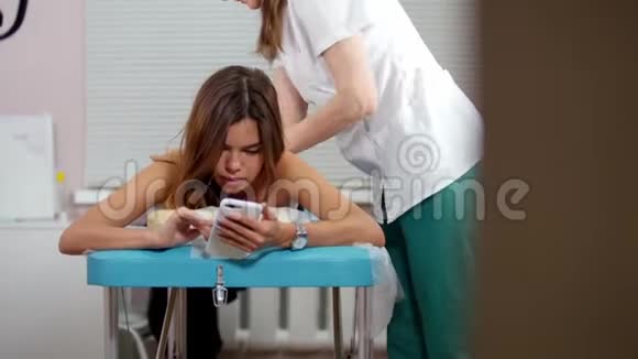 一位年轻女子接受放松的背部按摩在社交网络上坐着她的手机视频的预览图